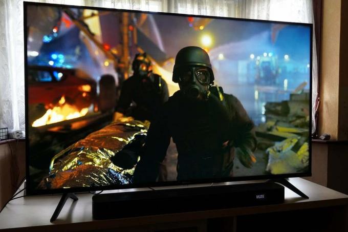 Tahta bir masanın üzerinde duran siyah bir Hisense A7200 TV, siyah takım elbiseli ve gaz maskeli bir ceset taşıyan iki adamı gösteriyor, Tenet'ten bir sahne